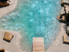 Vue aérienne d’un bassin de forme libre bordé de sable, traversé par un ponton de bois